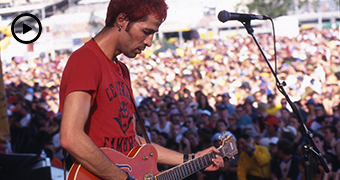 Before a blurred large crowd of people, a man in red T-shirt plays guitar, raised on a stage.