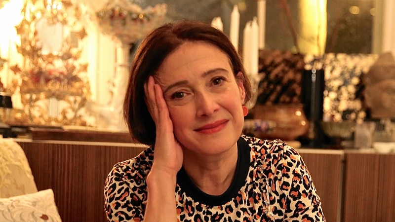 Woman with short hair and animal print shirt sits on armchair with pillows, with face leaning into hand.