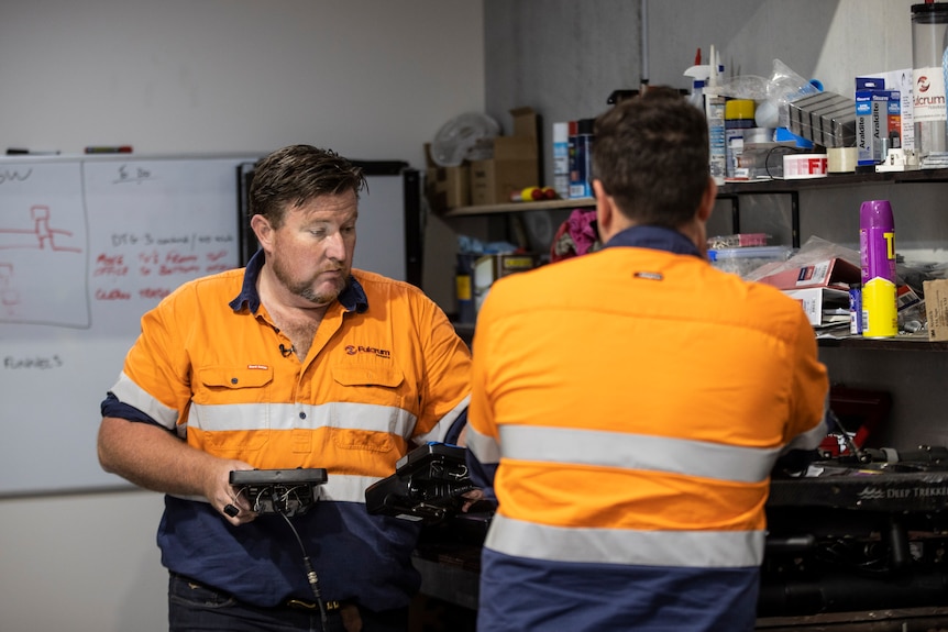 Two men in orange hi-visability shirts, one with his back to the camera. The other is bearded and holds an industrial robot.