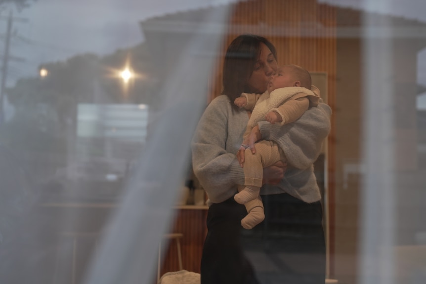 A middle aged white woman with brown hair is seen through a window holding a baby 