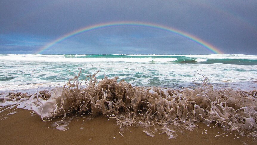 Wave crashes onto beach, rainbow in background