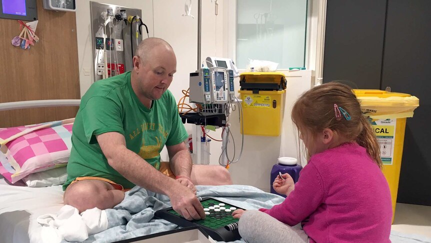 Jarrod Lyle sits on a hospital bed with his daughter Lusi playing a board game.