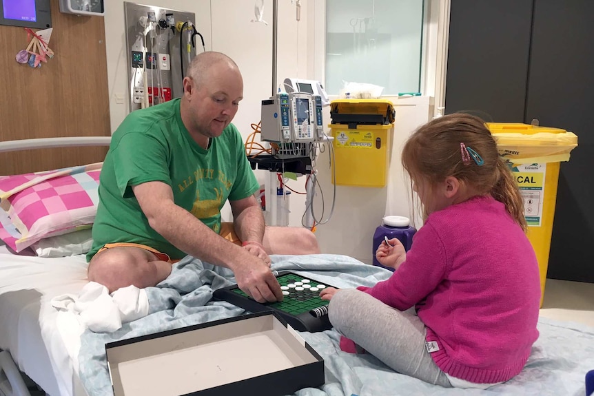 Jarrod Lyle sits on a hospital bed with his daughter Lusi playing a board game.