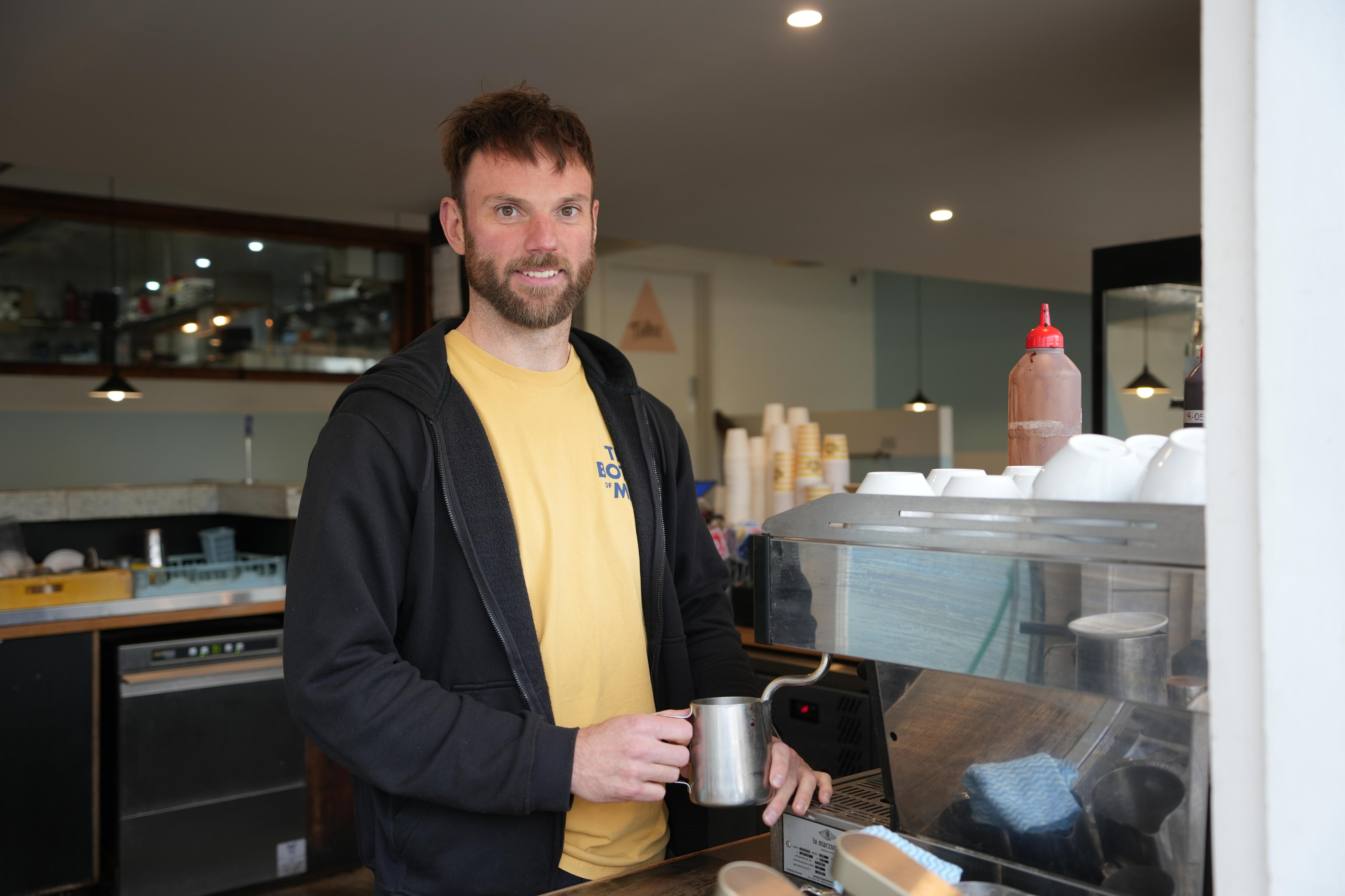 A man with brown hair in a yellow shirt and black jacket holds a silver jug under a milk frother on a coffee machine.