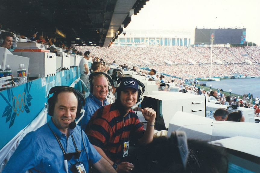 Three men with headsets sitting in the commentary section of a stadium looking at the camera.