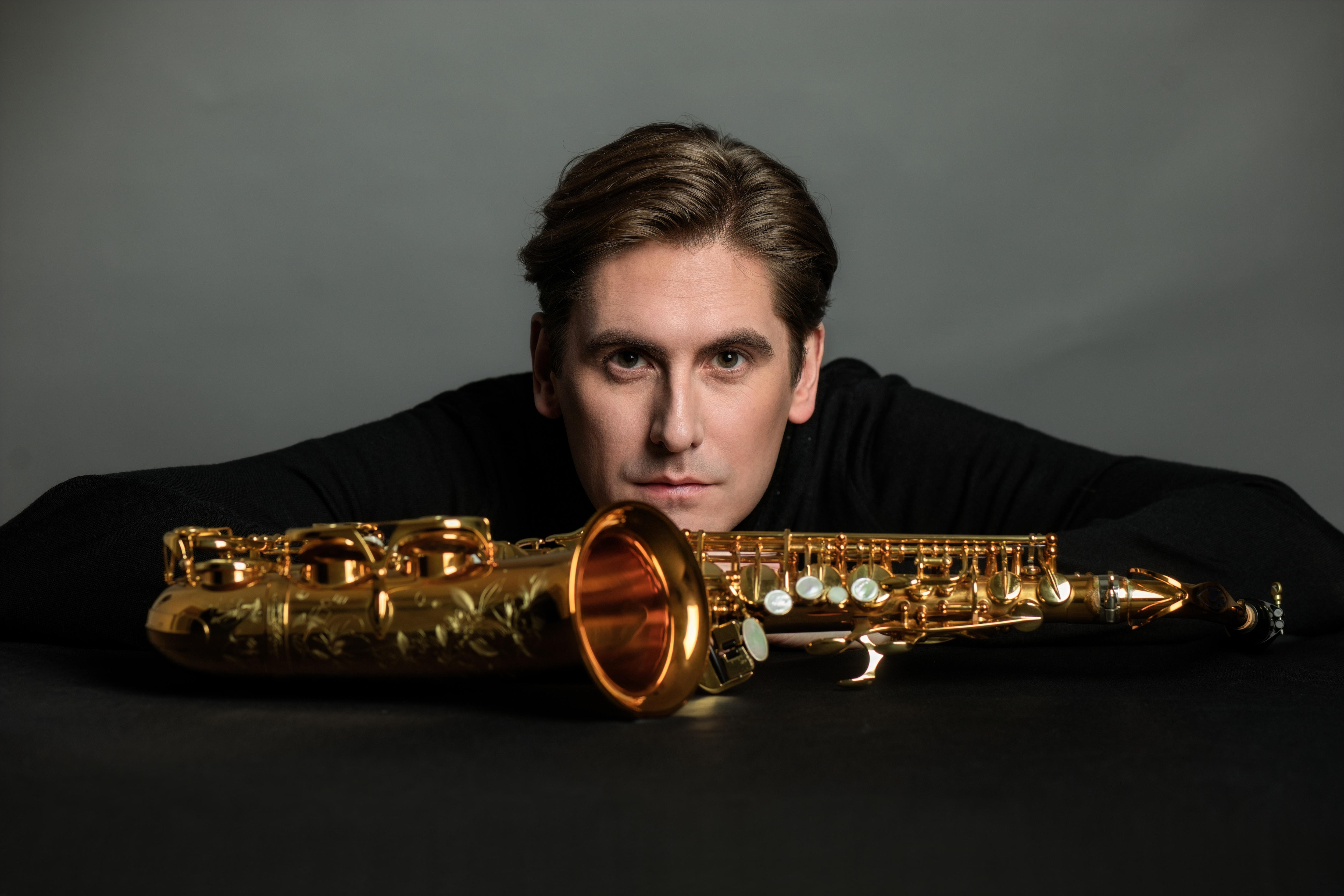 Saxophonist Jonathan Radford stares intently into the camera, his saxophone lying on its side in front of his face.