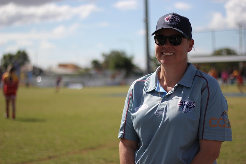 A woman in a polo shirt with a hat smiling and standing on a rugby pitch.