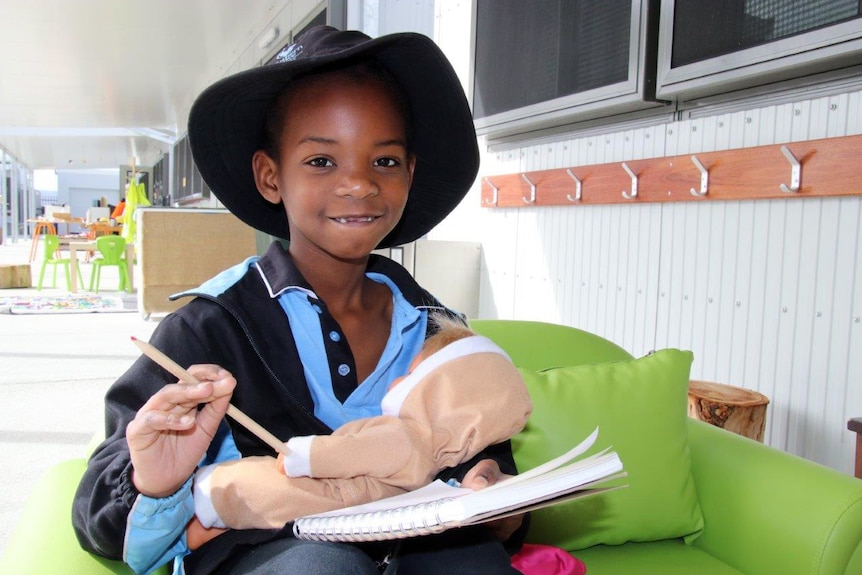 A student sits on a green lounge, holding a pencil, paper and a doll.