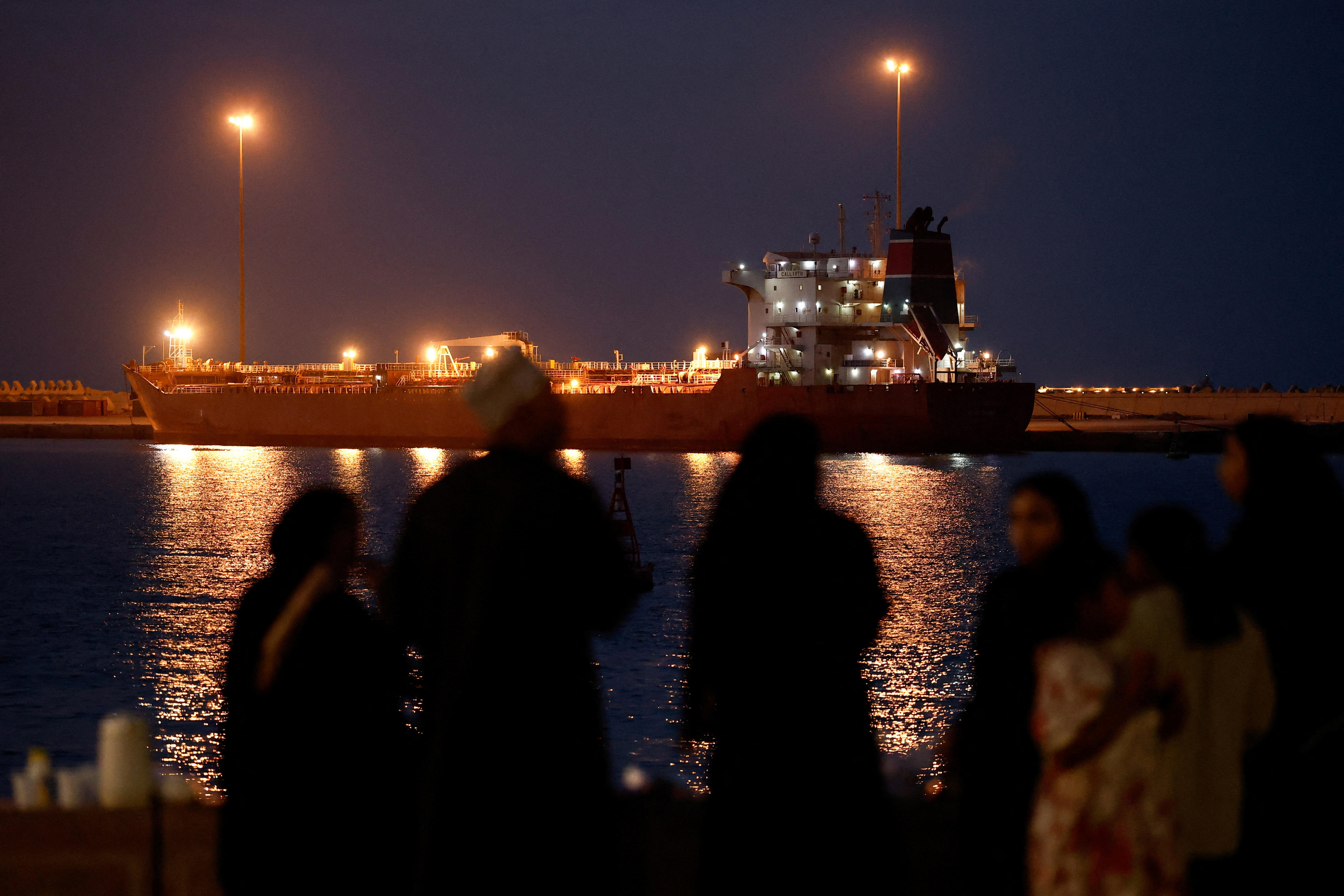 An oil tanker sits in a port at night, with silhouettes in front of it as people watch on.