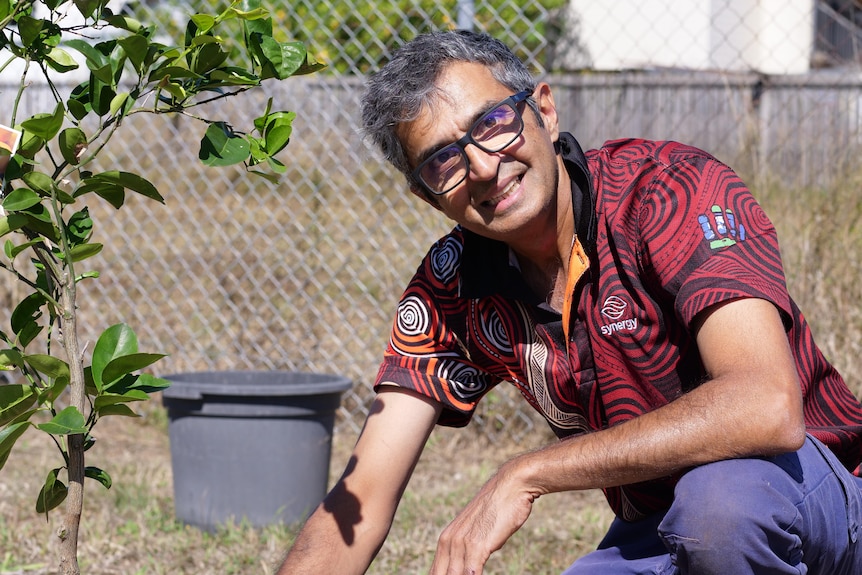 A man crouches beside a newly planted tree and smiles at the camera.