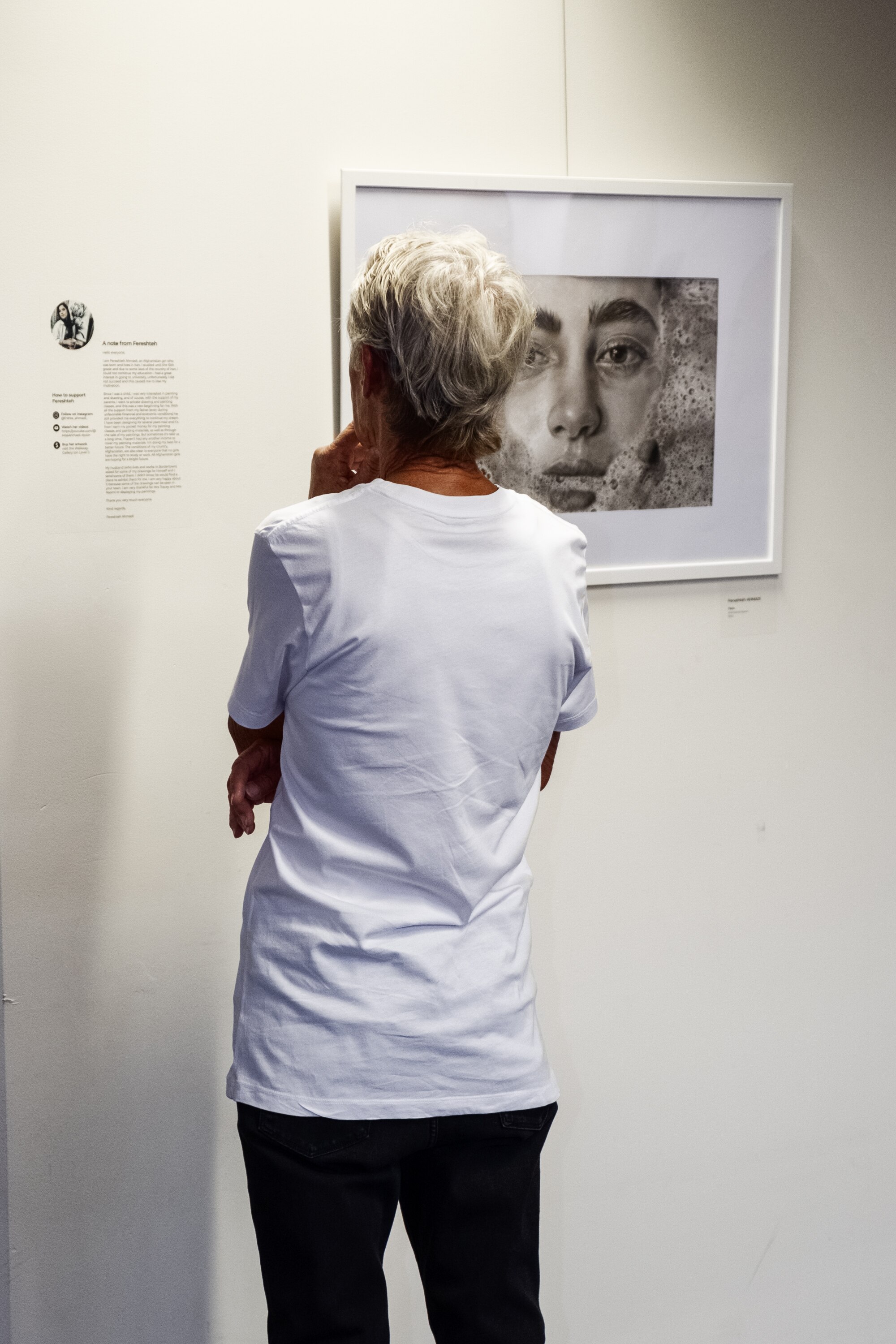 A woman stands in an art gallery, regarding a drawing hanging on the wall.