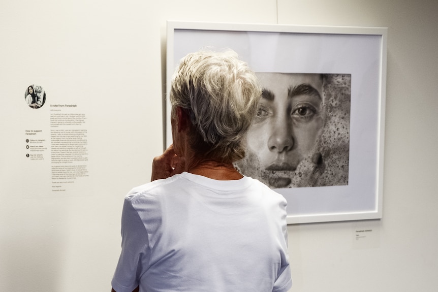 A woman stands in an art gallery, regarding a drawing hanging on the wall.