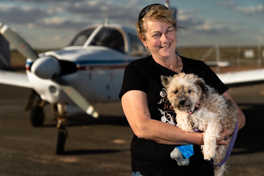 A woman holds a shih tzu and smiles while standing on an outback airport tarmac with a small plane in the background.