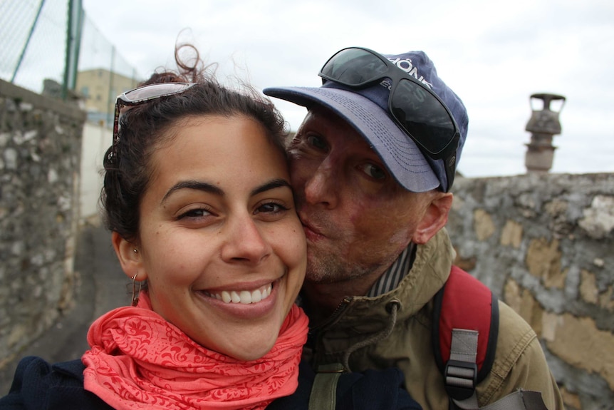 A man wearing a hat gives a kiss on a cheek to his smiling girlfriend