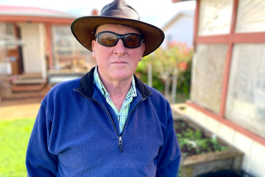 A middle-aged white man with low vision standing in a backyard garden. He is wearing glasses and a rimmed hat