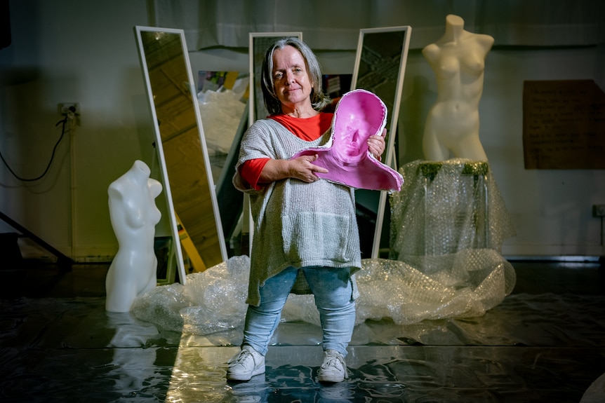 A woman living with dwarfism holding a pink body mould in front of a mirror