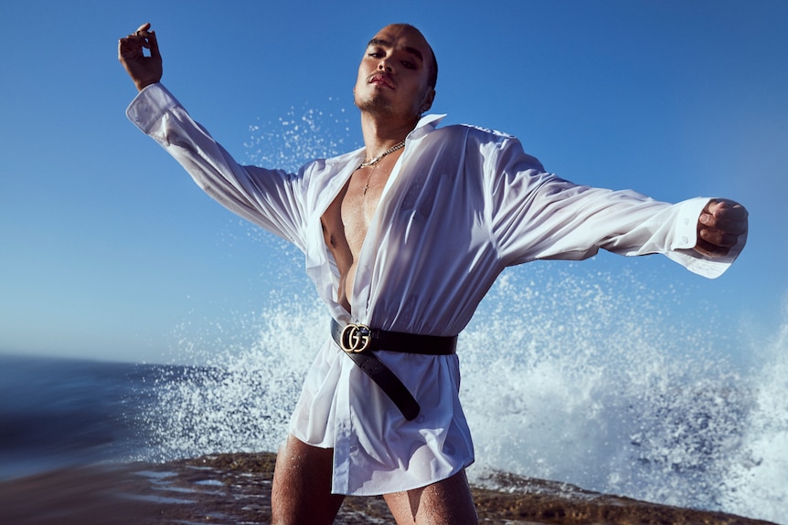 A man wearing a kimono stands on a rock with arms outstretched as a wave crashes behind him.