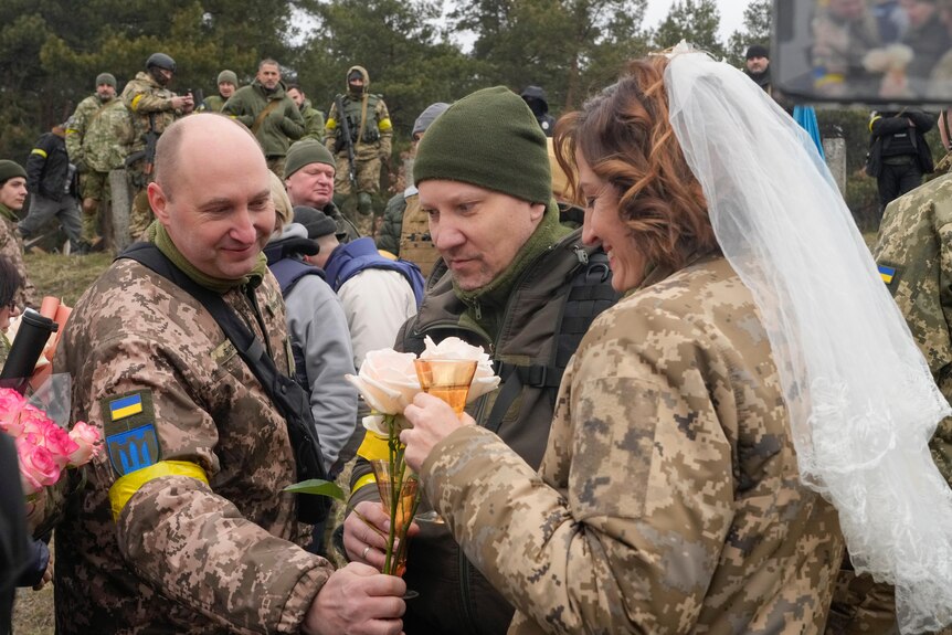 A man and woman in military uniforms hold flowers, the woman wearing a wedding veil, as other service people stand nearby.