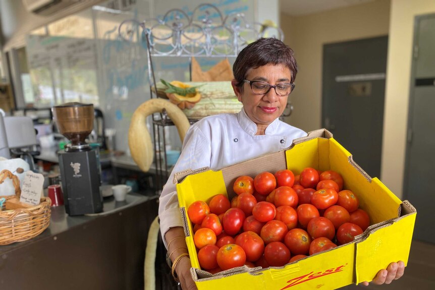 A woman stands in front of a cafe counter holding a cardboard box of tomatoes.