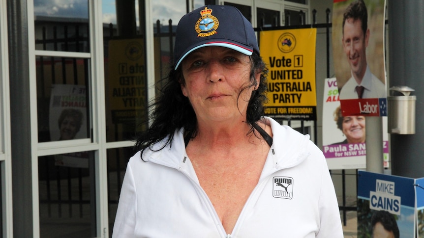 A woman in a white top stares at the camera in front of a United Australia Party poster