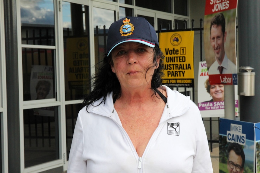 A woman in a white top stares at the camera in front of a United Australia Party poster