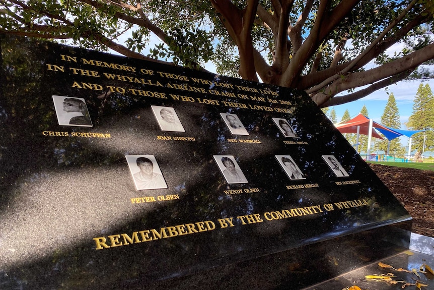 A memorial stone with eight pictures on it. It is in the shade of a large tree with a beach behind.