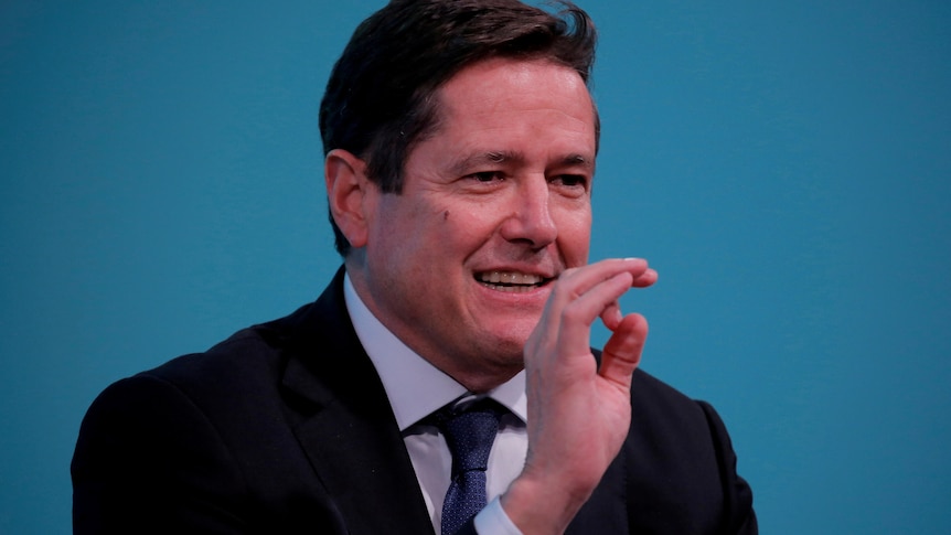 A man gestures as he speaks in front on a blue background.