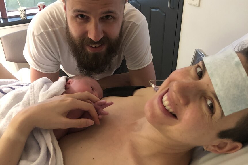 A woman in hospital with her baby on her chest, and her partner in the background