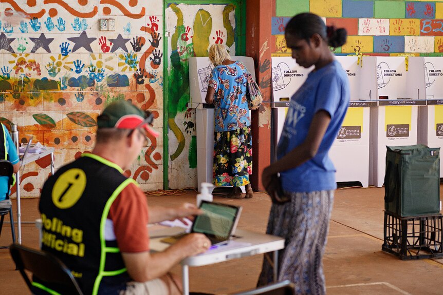 A man sits at a desk looking at a computer and a woman stands in front of him inside a voting centre.