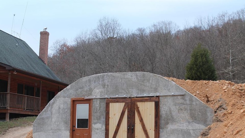 A grey bunker style room with barn doors covered in dirt with trees in the background.