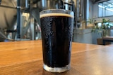 A full glass of dark beer sits on a table in a brewery.