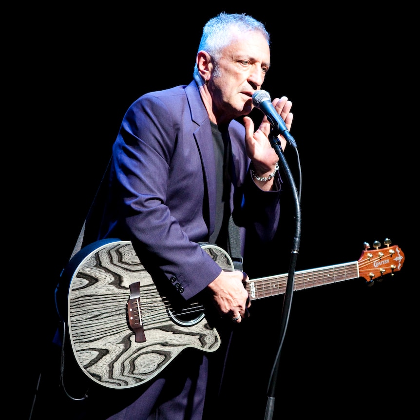 John Waters holds a guitar and speaks into a microphone