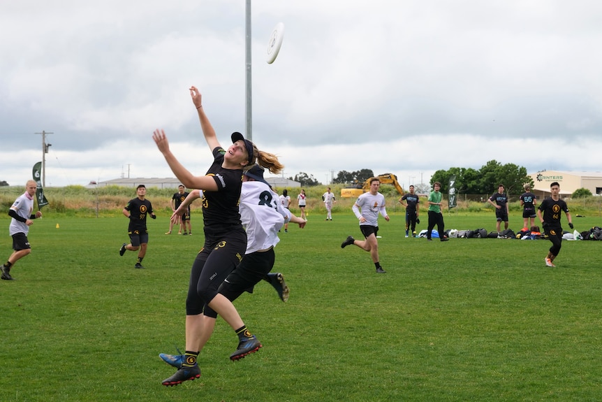 A woman in black leaps into the air trying to catch a Frisbee.