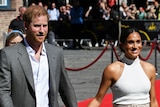 Prince harry in an open-collard shirt and grey jacket, holds hands with his wife Meghan in a beige halter neck