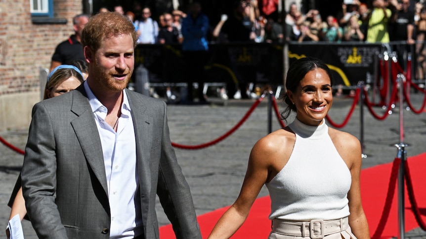 Prince harry in an open-collard shirt and grey jacket, holds hands with his wife Meghan in a beige halter neck