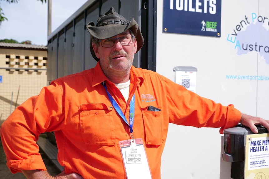 Arthur wearing a bright orange button up shirt, worn out kangaroo leather hat, portaloos behind.