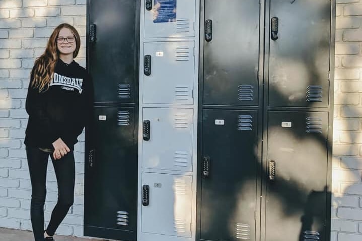 A young girl wearing black, stands in front of a set of lockers.