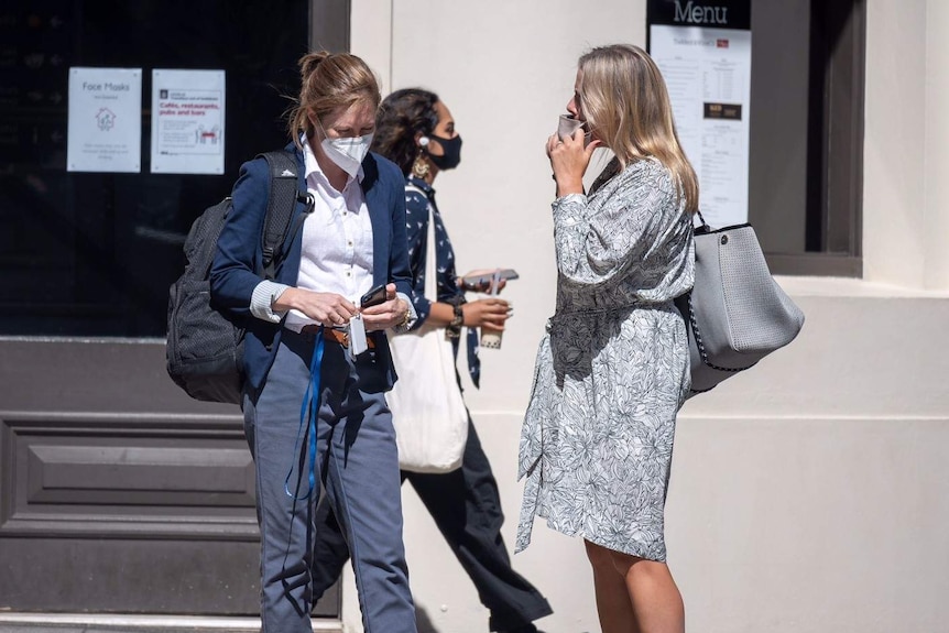 Two women stand talking to each other wearing masks as a third woman walks behind them.