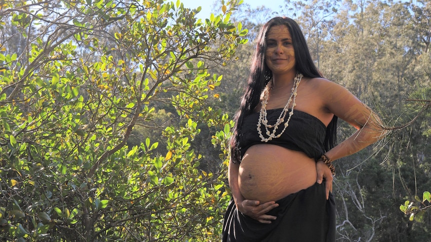 a pregnant Indigenous woman stands among trees