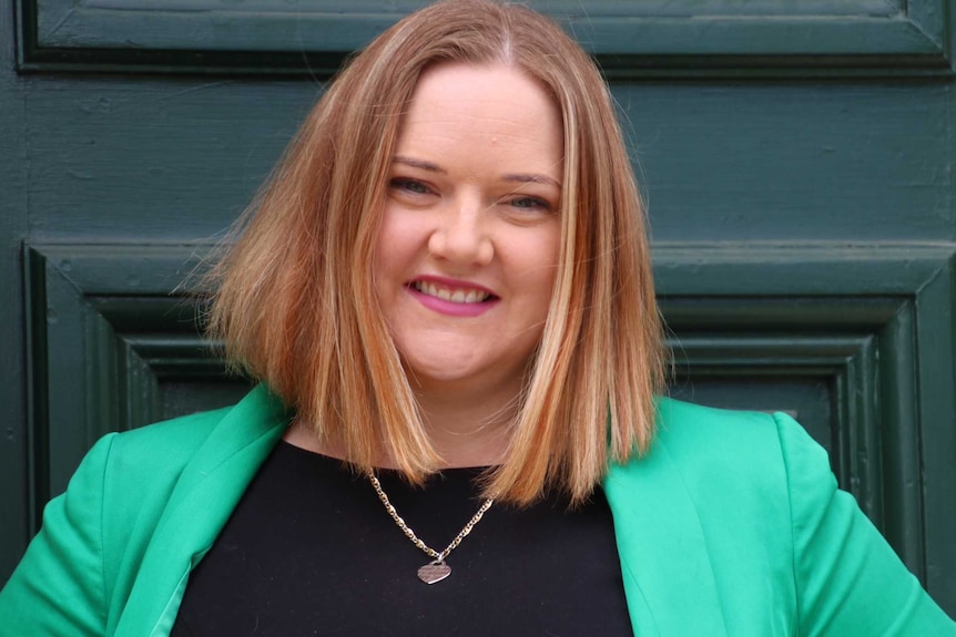 Jessica Stojkovski wearing a green jacket and black top in front of a black door.