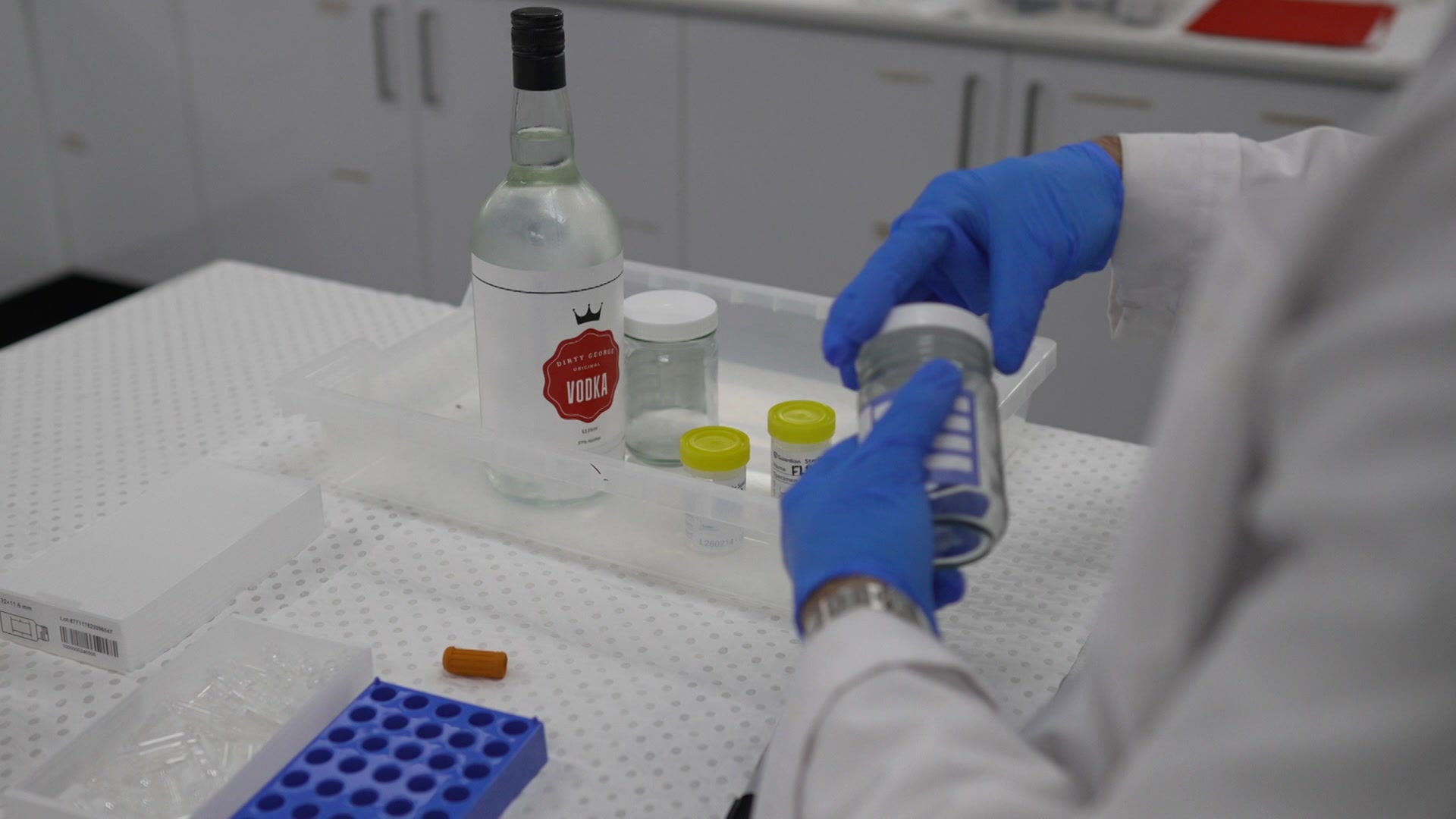 A person opening up a testing jar with a large bottle of alcohol sitting in a plastic tray on a table.