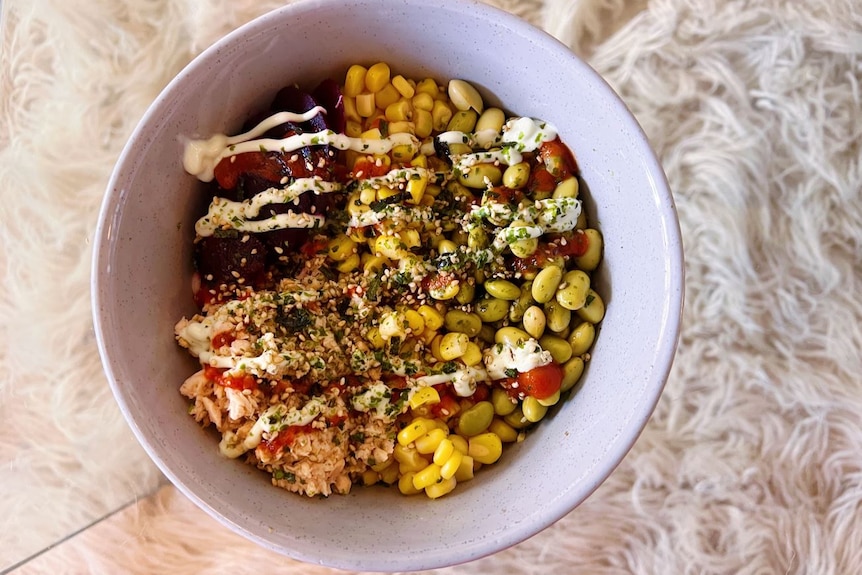 A grey speckled bowl is seen sitting atop a glass table with a white flokati rug underneath. The bowl is filled with a poke dish