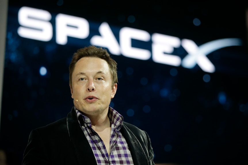 Elon Musk stands in front of glowing SpaceX sign.