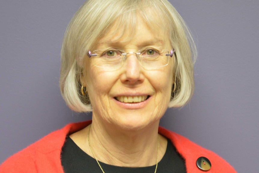 A woman wearing glasses, a red cardigan and a black knitted top smiles at the camera in front of a wall.