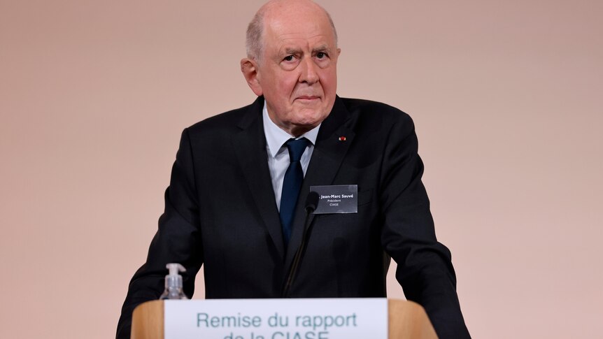 An old, balding man in a suit stands sternly at a lectern.