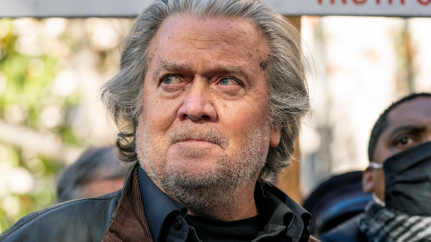 A white male with grey hair and stubble stares off camera