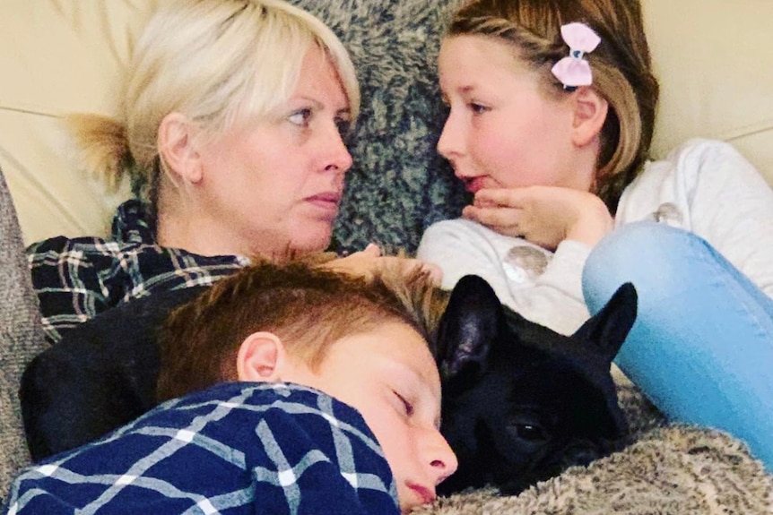 A woman and two kids relax on a bed with a black dog