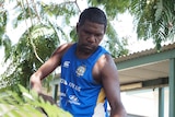 A young man in blue singlet lifts leaves with a rake in the shade of a tree