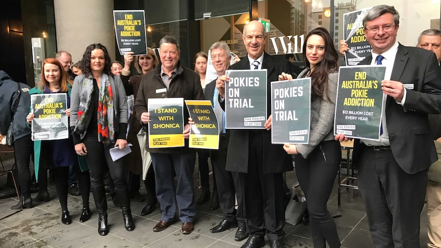 About a dozen supporters of the legal action hold up signs against the pokies.