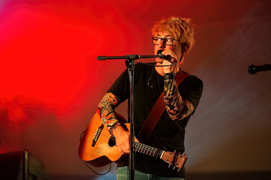 A man dressed as Ed Sheeran hp;ds a guitar and stands behind a microphone on stage. 